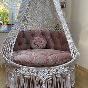 May include: A macrame swing chair with a canopy, featuring a dusty rose cushion and backrest. The chair has a round decorative pillow. The white macrame design includes tassels and fringe. The overall design creates a bohemian aesthetic.