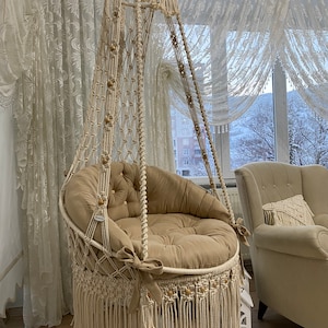 Può includere: Una sedia a dondolo in macramé beige con una frangia spessa e un cuscino beige. La sedia è appesa a un soffitto bianco e si trova in una stanza con tende bianche.