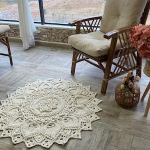 以下が含まれることがあります： クリーム色の、詳細な花柄の丸いかぎ針編みラグ。 ラグは、クリーム色のクッションが付いた木製の椅子の近くにある、明るい茶色の木の床に置かれています。 花の入った小さな柳のバスケットが近くにあります。