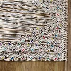 Puede incluir: Un tapiz de macram&eacute; blanco con un patr&oacute;n geom&eacute;trico. El macram&eacute; est&aacute; decorado con hilo de colores en varios tonos de rosa, azul, verde, amarillo y morado. El tapiz est&aacute; sujeto a una varilla de madera.