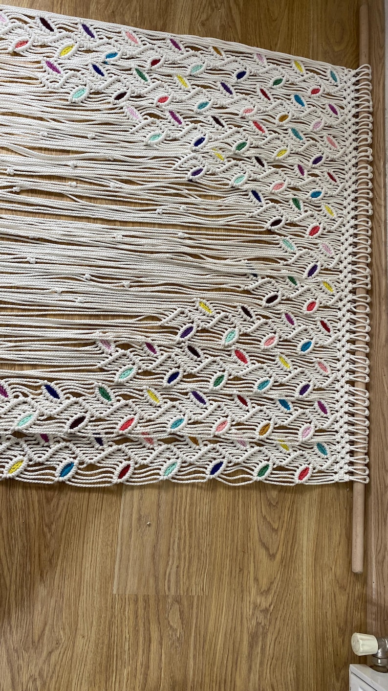 Puede incluir: Un tapiz de macram&eacute; blanco con un patr&oacute;n geom&eacute;trico. El macram&eacute; est&aacute; decorado con hilo de colores en varios tonos de rosa, azul, verde, amarillo y morado. El tapiz est&aacute; sujeto a una varilla de madera.