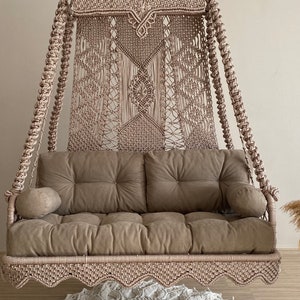 Può includere: Una sedia a dondolo in macramè beige con un cuscino marrone e due cuscini. La sedia è appesa al soffitto e si trova di fronte a un muro bianco. Un tappeto bianco all'uncinetto si trova sul pavimento sotto la sedia.