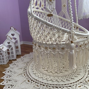 K&ouml;nnte beinhalten: Ein wei&szlig;er Makramee-H&auml;ngesessel mit Holzperlen und Quasten, der auf einem wei&szlig;en geh&auml;kelten Teppich mit Mandala-Muster steht.