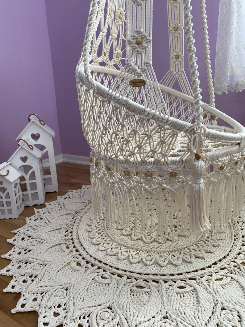 K&ouml;nnte beinhalten: Ein wei&szlig;er Makramee-H&auml;ngesessel mit Holzperlen und Quasten, der auf einem wei&szlig;en geh&auml;kelten Teppich mit Mandala-Muster steht.