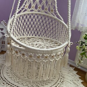 K&ouml;nnte beinhalten: Ein wei&szlig;er Makramee-H&auml;ngesessel mit Holzperlen und Fransen. Der Sessel h&auml;ngt an einer lila Wand und steht auf einem wei&szlig;en geh&auml;kelten Teppich.