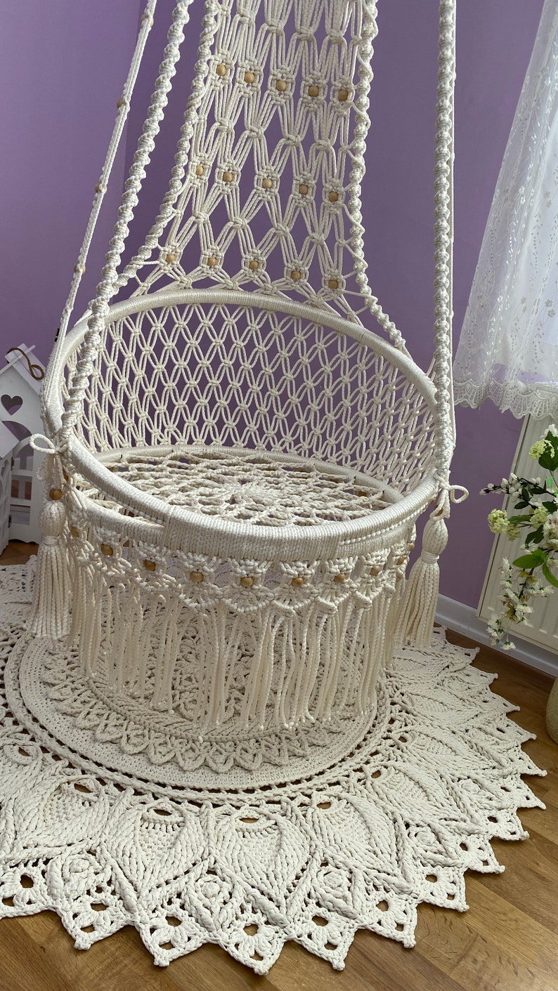 K&ouml;nnte beinhalten: Ein wei&szlig;er Makramee-H&auml;ngesessel mit Holzperlen und Fransen. Der Sessel h&auml;ngt an einer lila Wand und steht auf einem wei&szlig;en geh&auml;kelten Teppich.