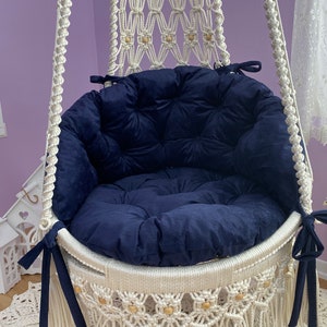 K&ouml;nnte beinhalten: Ein wei&szlig;er Makramee-Schaukelstuhl mit einem marineblauen Kissen. Der Stuhl hat Holzperlen und Fransen.