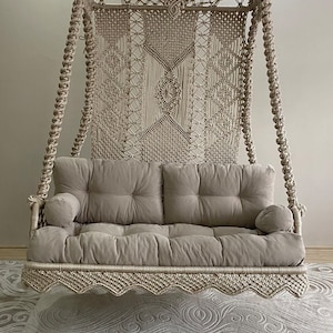 May include: A beige macrame swing chair with a thick rope design and a beige cushion. The swing chair is hanging from the ceiling.