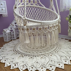 K&ouml;nnte beinhalten: Ein wei&szlig;er Makramee-H&auml;ngesessel mit Holzperlen und Quasten, der auf einem wei&szlig;en Makramee-Teppich mit kreisf&ouml;rmigem Muster steht.