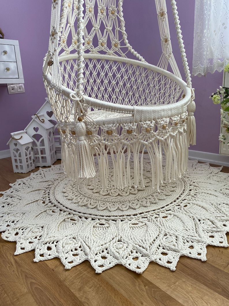K&ouml;nnte beinhalten: Ein wei&szlig;er Makramee-H&auml;ngesessel mit Holzperlen und Quasten, der auf einem wei&szlig;en Makramee-Teppich mit kreisf&ouml;rmigem Muster steht.