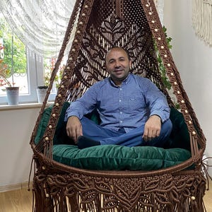 Peut inclure: Un fauteuil suspendu en macramé marron avec un coussin vert et des franges. La chaise est ornée de perles de bois et est suspendue au plafond. La personne est assise dans la chaise.