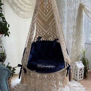 Op de afbeelding: Een crèmekleurige macramé hangstoel met een blauw fluwelen kussen. De stoel hangt aan een houten haak aan het plafond en heeft een franje rand. De stoel staat op een crèmekleurig gehaakt tapijt.