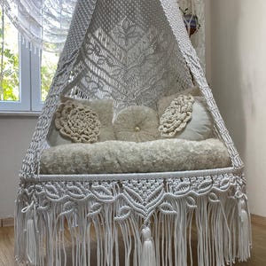 May include: White macrame hanging chair with a fringe border and a tassel. The chair is decorated with white pillows and is hanging in a room with white curtains.