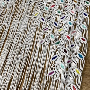 Puede incluir: Un tapiz de macram&eacute; blanco con un patr&oacute;n geom&eacute;trico y acentos coloridos. El tapiz est&aacute; hecho de cord&oacute;n de algod&oacute;n natural y presenta una variedad de nudos y bucles.