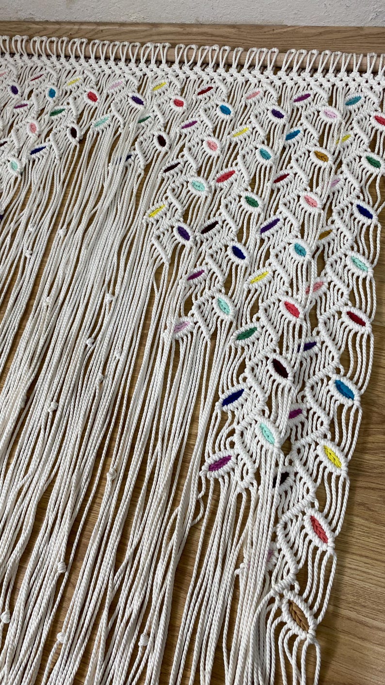 Puede incluir: Un tapiz de macram&eacute; blanco con un patr&oacute;n geom&eacute;trico y acentos coloridos. El tapiz est&aacute; hecho de cord&oacute;n de algod&oacute;n natural y presenta una variedad de nudos y bucles.