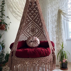 May include: A macrame hanging chair with a burgundy cushion and a pink pillow. The chair is hanging from the ceiling and has a fringe around the bottom.
