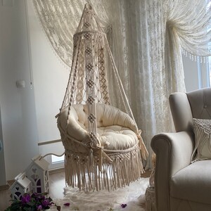 May include: A white macrame hanging chair with a white cushion and fringe. The chair is hanging from the ceiling in a room with white curtains.