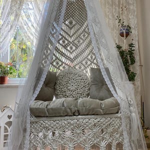 May include: A white macrame hammock chair with a white lace canopy and a beige cushion. The chair is hanging from the ceiling and has a fringe trim.