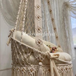 May include: A cream-coloured macrame swing chair with a white cushion and tassels. The chair is hanging from the ceiling and has a wooden bead detail.
