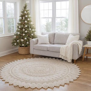 May include: A cream-coloured, round, crocheted rug with a detailed floral pattern. The rug is placed on a hardwood floor in a living room setting. A decorated Christmas tree and a light grey sofa are in the background.