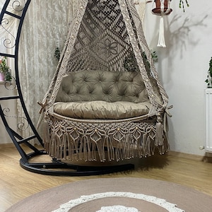 May include: A beige macrame hanging chair with a brown cushion and fringe. The chair is suspended from a black metal stand and is hanging over a brown rug with a white crocheted design.