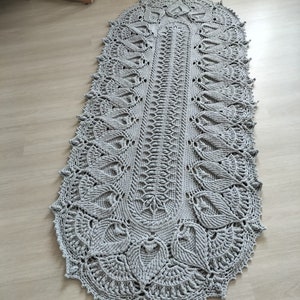 May include: A gray crocheted oval rug with a delicate floral pattern. The rug has a fringed edge and is made with a thick yarn.