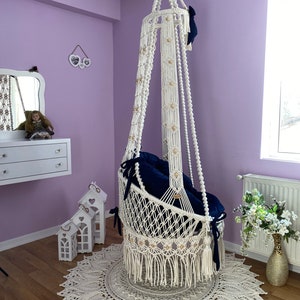 K&ouml;nnte beinhalten: Ein wei&szlig;er Makramee-H&auml;ngesessel mit einem marineblauen Kissen. Der Sessel h&auml;ngt von der Decke in einem Raum mit violetten W&auml;nden. Der Sessel steht auf einem wei&szlig;en geh&auml;kelten Teppich.