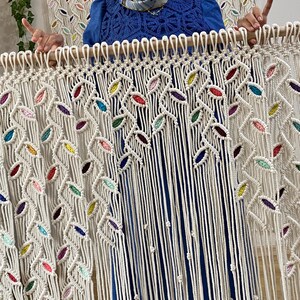 Puede incluir: Un tapiz de macram&eacute; blanco con detalles coloridos. El tapiz presenta un patr&oacute;n de hojas repetido y est&aacute; hecho con cord&oacute;n de algod&oacute;n natural.