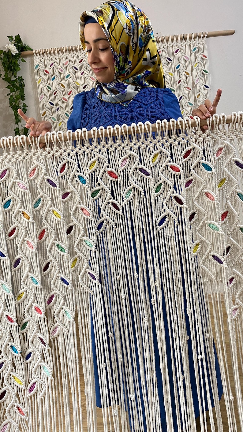 Puede incluir: Un tapiz de macram&eacute; blanco con detalles coloridos. El tapiz presenta un patr&oacute;n de hojas repetido y est&aacute; hecho con cord&oacute;n de algod&oacute;n natural.