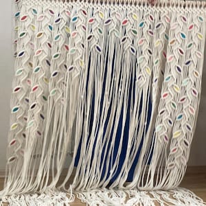 Puede incluir: Un tapiz de macram&eacute; blanco con detalles coloridos. El tapiz est&aacute; hecho de cuerdas anudadas y presenta un patr&oacute;n de hojas.