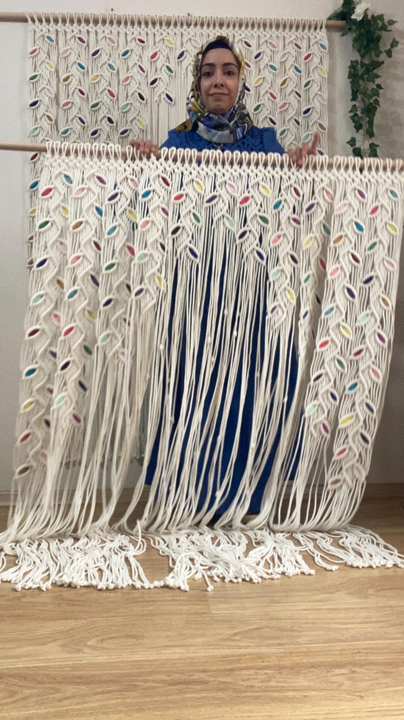 Puede incluir: Un tapiz de macram&eacute; blanco con detalles coloridos. El tapiz est&aacute; hecho de cuerdas anudadas y presenta un patr&oacute;n de hojas.