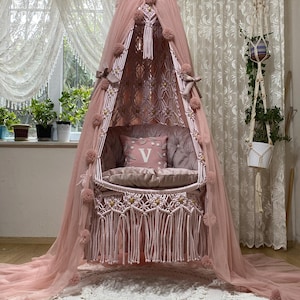 Peut inclure: Un fauteuil suspendu en macramé rose avec un coussin blanc et un baldaquin rose. Le fauteuil est suspendu à un crochet de plafond et est entouré d'un tapis crocheté blanc.