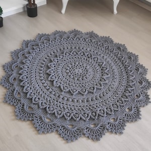 Könnte beinhalten: Ein runder, grauer Häkelteppich mit einem zarten Spitzenmuster. Der Teppich hat mehrere Schichten mit komplizierten Designs, die eine optisch ansprechende und strukturierte Oberfläche schaffen.