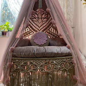 May include: A handcrafted macrame swing bed with a pink canopy and decorative fringe. The bed features a round, floral-shaped pillow and several other pillows. The canopy is draped with pink tulle and adorned with pink pom-poms. The bed is suspended from the ceiling.