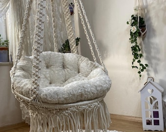 Könnte beinhalten: Ein weißer Makramee-Hängesessel mit einem weißen Kissen und Fransen. Der Sessel hängt von der Decke in einem Raum mit einer weißen Wand und einem weißen Teppich mit Blumenmuster.