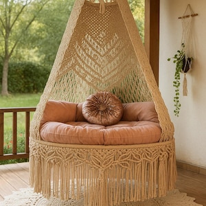 Peut inclure: Un fauteuil suspendu en macramé beige avec une base ronde à franges et un sommet conique. Le fauteuil comprend un coussin rond rose blush et des coussins décoratifs. Un tapis rond crème est sous la chaise, avec une plante suspendue en arrière-plan.