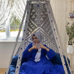K&ouml;nnte beinhalten: Eine Frau in einem blauen Kleid und einem hellgrauen Kopftuch sitzt in einem wei&szlig;en Makramee-H&auml;ngesessel mit blauen Perlen. Der Sessel hat ein blaues Kissen und eine Fransen an der Unterseite.