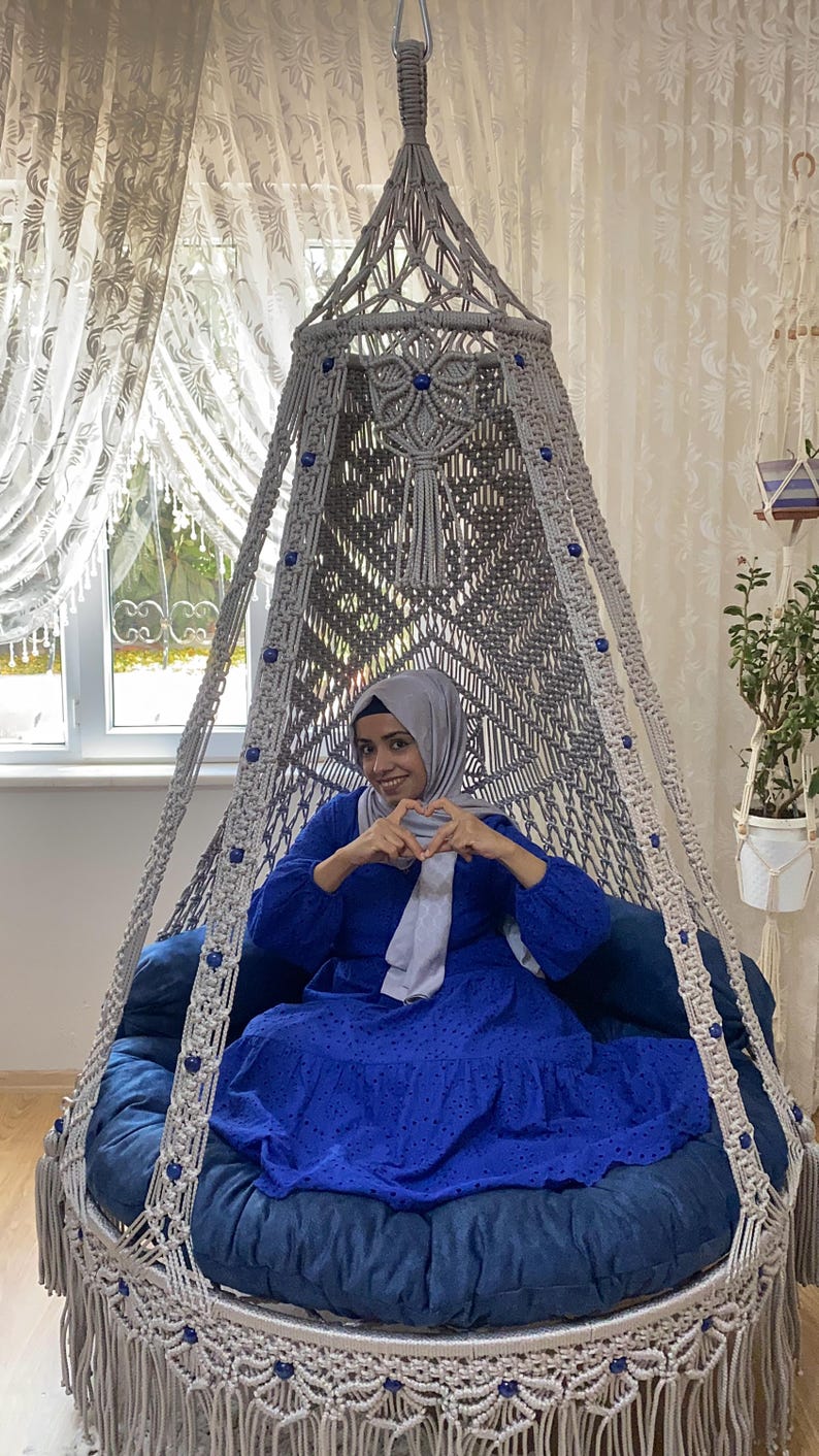 K&ouml;nnte beinhalten: Eine Frau in einem blauen Kleid und einem hellgrauen Kopftuch sitzt in einem wei&szlig;en Makramee-H&auml;ngesessel mit blauen Perlen. Der Sessel hat ein blaues Kissen und eine Fransen an der Unterseite.