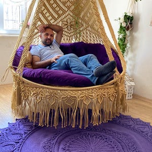 May include: A large, round, macrame hanging chair with a fringe border. The chair is made of natural-colored cord and is hanging from the ceiling. A person is sitting in the chair, which is resting on a purple crocheted rug.