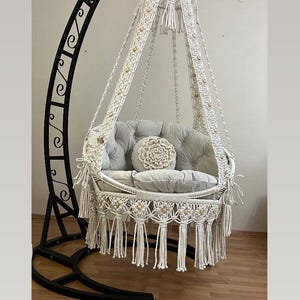 May include: A white macrame hanging chair with a gray cushion and a white pillow. The chair is suspended from a black metal stand.