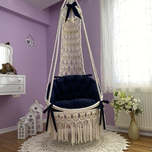 K&ouml;nnte beinhalten: Ein wei&szlig;er Makramee-H&auml;ngesessel mit einem dunkelblauen Kissen und schwarzen Schleifenakzenten. Der Sessel h&auml;ngt von der Decke in einem Raum mit einem wei&szlig;en Teppich und einer Blumenvase.