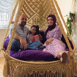 Può includere: Una famiglia di tre persone è seduta su una grande sedia a dondolo rotonda in macramè con un cuscino viola. La sedia è realizzata in corda naturale e ha un bordo a frangia. La sedia è appesa al soffitto.