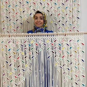 May include: A white macrame wall hanging with colorful accents. The hanging features a leaf pattern and is made with thick cotton cord.
