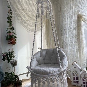 Può includere: Poltrona sospesa in macramé bianco con un cuscino bianco e frange. La poltrona è appesa a un gancio da soffitto bianco e si trova in una stanza con tende bianche.
