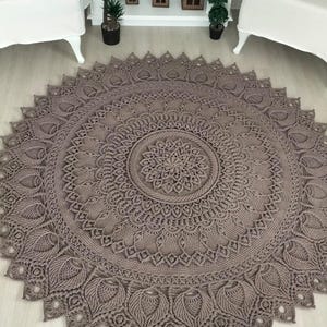 Könnte beinhalten: Ein großer, runder, aufwendig gehäkelter Teppich in einem gedämpften Taupe-Ton. Der Teppich hat ein detailliertes Mandala-Design mit einem zentralen Blumenmotiv und einem gewellten Rand. Der Teppich liegt auf einem hellen Holzboden.