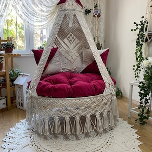 May include: A white macrame hanging chair with a red cushion and a white macrame rug underneath. The chair has a fringe border and a red bow at the top.