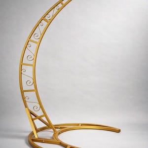 May include: A gold-colored, crescent-shaped metal stand with decorative scrollwork. The stand has a circular base and a curved top with a hook, likely for hanging items. The design is ornate and suitable for events or decor.