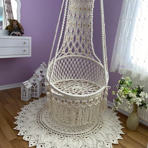 K&ouml;nnte beinhalten: Ein wei&szlig;er Makramee-H&auml;ngesessel mit Fransenbord&uuml;re und ein wei&szlig;er geh&auml;kelter Teppich mit Mandala-Muster.