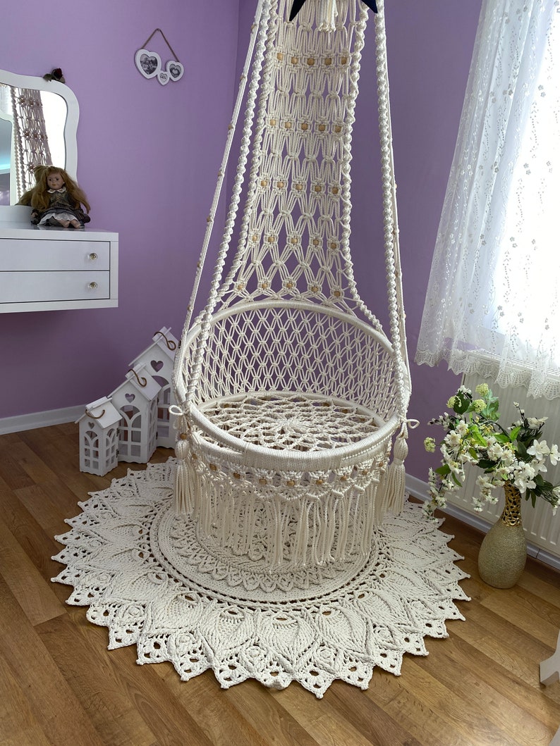 K&ouml;nnte beinhalten: Ein wei&szlig;er Makramee-H&auml;ngesessel mit Fransenbord&uuml;re und ein wei&szlig;er geh&auml;kelter Teppich mit Mandala-Muster.