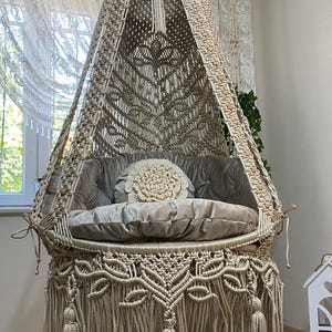 May include: A beige macrame hanging chair with a soft, gray cushion and a decorative, round pillow. The chair features intricate knotwork, tassels, and wooden beads. The chair is suspended from the ceiling, creating a cozy and stylish seating area.